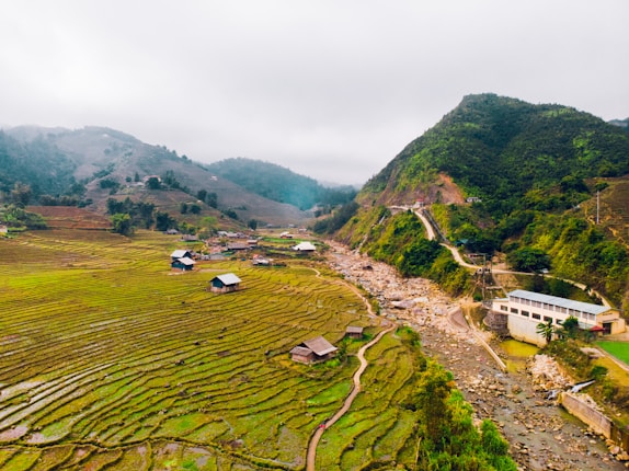 Terraced rice fields stretch across the landscape, interspersed with small houses. A winding path leads through the fields alongside a rocky riverbed. Lush green hills rise in the background, partially covered by mist.