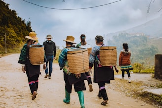 A group of people walk along a dirt path in a rural setting, each carrying large woven baskets on their backs. They are dressed in traditional, colorful attire with hats, and the sky is overcast. Lush green hills and scattered houses are visible in the background, depicting a mountainous landscape.