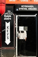 A storefront with signage advertising keyboards, synths, and organs. A person in a white shirt and black pants is standing inside the door with a sign that reads 'POSH' facing outwards. The exterior features graffiti and an arrow pointing downstairs labeled 'Keys'. The color scheme includes a bright red awning with words 'New', 'Used', and 'Vinyl'.