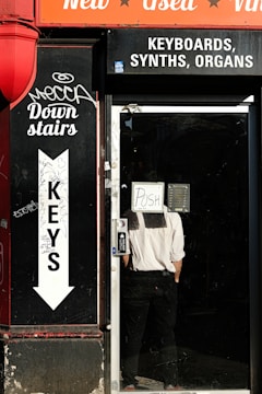 A storefront with signage advertising keyboards, synths, and organs. A person in a white shirt and black pants is standing inside the door with a sign that reads 'POSH' facing outwards. The exterior features graffiti and an arrow pointing downstairs labeled 'Keys'. The color scheme includes a bright red awning with words 'New', 'Used', and 'Vinyl'.