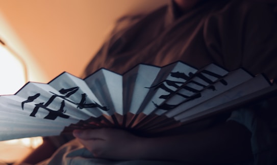 A person is holding a traditional hand fan with Japanese calligraphy written on it. The setting appears to be dimly lit, creating a soft and intimate atmosphere. The person is wearing a garment that resembles traditional Japanese clothing.