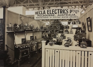 A vintage display of various electric heating appliances showcased in a booth with a prominent sign that reads 'Hecla Electrics Pty. Ltd. Melbourne, Australia.' The booth is filled with numerous appliances such as stoves, toasters, and kettles, neatly arranged on shelves and stands. The display is organized and packed, giving a sense of a classic trade show environment.