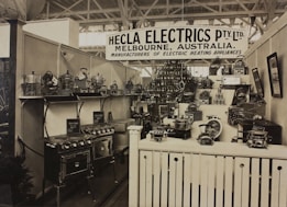 A vintage display of various electric heating appliances showcased in a booth with a prominent sign that reads 'Hecla Electrics Pty. Ltd. Melbourne, Australia.' The booth is filled with numerous appliances such as stoves, toasters, and kettles, neatly arranged on shelves and stands. The display is organized and packed, giving a sense of a classic trade show environment.