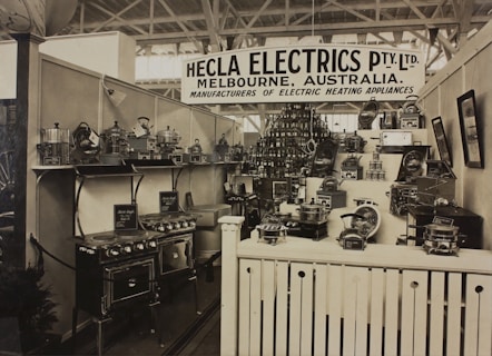 A vintage display of various electric heating appliances showcased in a booth with a prominent sign that reads 'Hecla Electrics Pty. Ltd. Melbourne, Australia.' The booth is filled with numerous appliances such as stoves, toasters, and kettles, neatly arranged on shelves and stands. The display is organized and packed, giving a sense of a classic trade show environment.