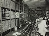A large, organized warehouse filled with shelves stacked with boxes and electronic parts. Several workers, some wearing lab coats, are engaged in tasks like assembling items. The floor is lined with rows of fans or similar objects.