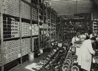 A large, organized warehouse filled with shelves stacked with boxes and electronic parts. Several workers, some wearing lab coats, are engaged in tasks like assembling items. The floor is lined with rows of fans or similar objects.