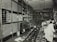 A large, organized warehouse filled with shelves stacked with boxes and electronic parts. Several workers, some wearing lab coats, are engaged in tasks like assembling items. The floor is lined with rows of fans or similar objects.