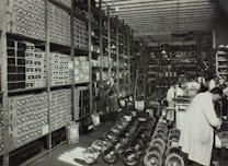A large, organized warehouse filled with shelves stacked with boxes and electronic parts. Several workers, some wearing lab coats, are engaged in tasks like assembling items. The floor is lined with rows of fans or similar objects.