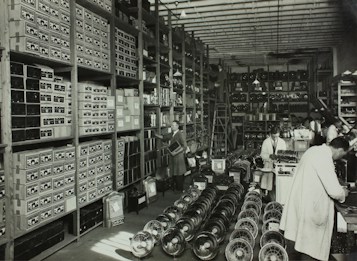 A large, organized warehouse filled with shelves stacked with boxes and electronic parts. Several workers, some wearing lab coats, are engaged in tasks like assembling items. The floor is lined with rows of fans or similar objects.