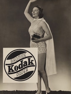 A woman is standing and smiling while wearing a vintage-style swimsuit and holding a small camera. She waves with her other hand. A cloak is draped over her shoulders. The image has a sepia tone and features a prominent Kodak advertisement in the foreground.