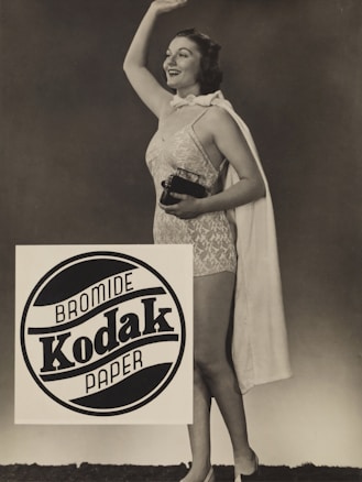 A woman is standing and smiling while wearing a vintage-style swimsuit and holding a small camera. She waves with her other hand. A cloak is draped over her shoulders. The image has a sepia tone and features a prominent Kodak advertisement in the foreground.