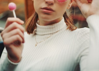 woman in white turtleneck sweater wearing red framed eyeglasses holding pink lollipop