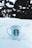 A white ceramic mug with a logo featuring a stylized figure is placed on a blanket of snow. The background shows blurred trees, indicating an outdoor setting with snow covering the ground.