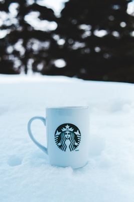 A white ceramic mug with a logo featuring a stylized figure is placed on a blanket of snow. The background shows blurred trees, indicating an outdoor setting with snow covering the ground.