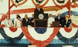 A group of people seated and standing on a stage decorated with red, white, and blue bunting. A man stands at a podium with the Presidential seal while others sit on either side, some in formal attire and others in naval uniforms. A banner with the word 'ANOGRAPHER' hangs behind them.