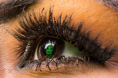 A close-up of an eye with elaborate eyelashes and makeup, reflecting a green theatrical mask with black accents. The eyelashes are long and curled, and the eye's iris is clearly visible, contrasting with the reflection in the pupil.