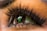 A close-up of an eye with elaborate eyelashes and makeup, reflecting a green theatrical mask with black accents. The eyelashes are long and curled, and the eye's iris is clearly visible, contrasting with the reflection in the pupil.