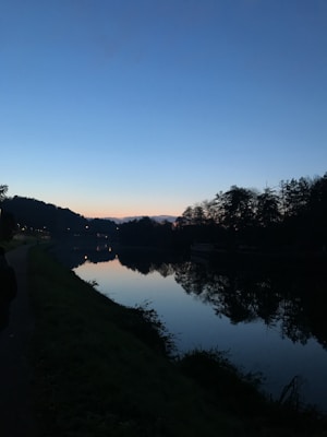 The image features a serene riverside scene during twilight. The sky transitions from a deep blue to a subtle orange hue near the horizon, creating a calm gradient effect. Tall trees line the riverbank, their silhouettes reflected crisply in the still water below. A dimly lit pathway follows the river, hinting at human presence without disrupting the tranquility of the natural setting.
