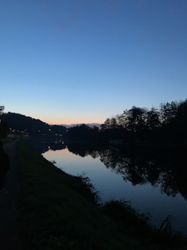 The image features a serene riverside scene during twilight. The sky transitions from a deep blue to a subtle orange hue near the horizon, creating a calm gradient effect. Tall trees line the riverbank, their silhouettes reflected crisply in the still water below. A dimly lit pathway follows the river, hinting at human presence without disrupting the tranquility of the natural setting.