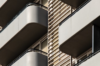 Modern architectural design featuring multiple balconies with smooth, curved edges. The structure incorporates horizontal slats and railings, providing a sleek, geometric appearance. Neutral tones enhance the minimalistic and sophisticated aesthetic.