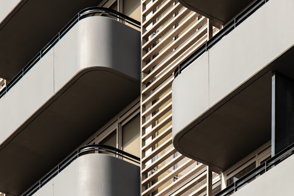 Modern architectural design featuring multiple balconies with smooth, curved edges. The structure incorporates horizontal slats and railings, providing a sleek, geometric appearance. Neutral tones enhance the minimalistic and sophisticated aesthetic.