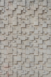 A textured wall composed of interlocking rectangular and square blocks, creating a three-dimensional pattern. The surface has a rough and concrete-like appearance.