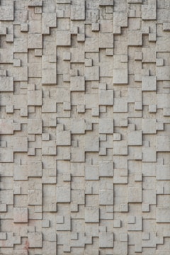 A textured wall composed of interlocking rectangular and square blocks, creating a three-dimensional pattern. The surface has a rough and concrete-like appearance.