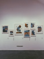 A collection of framed abstract artworks is displayed on a white wall. The pieces are characterized by geometric shapes and a combination of blue and earth tones, with each artwork differing slightly in composition and color balance.