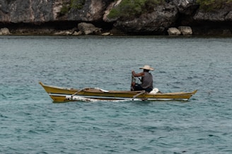 A person is seated in a small yellow and blue boat, navigating through clear, turquoise waters. The individual is wearing a wide-brimmed hat and appears to be operating some fishing equipment. Rocky cliffs with green foliage are visible in the background, providing a natural and rugged coastline.