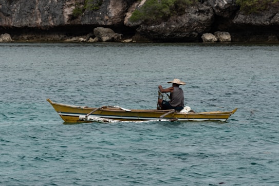 A person is seated in a small yellow and blue boat, navigating through clear, turquoise waters. The individual is wearing a wide-brimmed hat and appears to be operating some fishing equipment. Rocky cliffs with green foliage are visible in the background, providing a natural and rugged coastline.