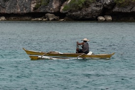 A person is seated in a small yellow and blue boat, navigating through clear, turquoise waters. The individual is wearing a wide-brimmed hat and appears to be operating some fishing equipment. Rocky cliffs with green foliage are visible in the background, providing a natural and rugged coastline.