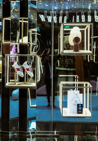 A luxurious retail display with illuminated shelves showcases a pair of gray shoes with red and blue stripes, a white knit hat on a mannequin stand, and a branded shopping bag with a small box. The reflective surfaces create a sophisticated and modern aesthetic.