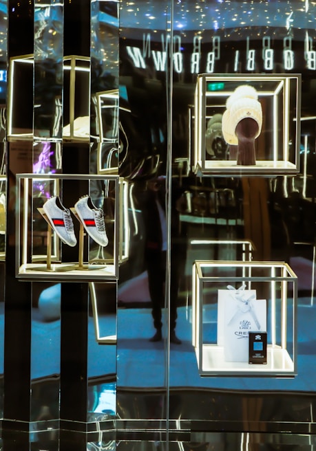 A luxurious retail display with illuminated shelves showcases a pair of gray shoes with red and blue stripes, a white knit hat on a mannequin stand, and a branded shopping bag with a small box. The reflective surfaces create a sophisticated and modern aesthetic.