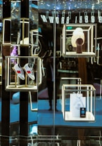 A luxurious retail display with illuminated shelves showcases a pair of gray shoes with red and blue stripes, a white knit hat on a mannequin stand, and a branded shopping bag with a small box. The reflective surfaces create a sophisticated and modern aesthetic.