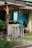 A rustic outdoor setup features a small wooden booth with a canopy, accompanied by two tall wooden stools painted in vibrant colors. The setting includes a gravel and grass ground with some lush greenery in the background. A sign with handwriting hangs on the booth, and there are decorative wheels and pots nearby.