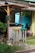A rustic outdoor setup features a small wooden booth with a canopy, accompanied by two tall wooden stools painted in vibrant colors. The setting includes a gravel and grass ground with some lush greenery in the background. A sign with handwriting hangs on the booth, and there are decorative wheels and pots nearby.