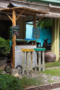 A rustic outdoor setup features a small wooden booth with a canopy, accompanied by two tall wooden stools painted in vibrant colors. The setting includes a gravel and grass ground with some lush greenery in the background. A sign with handwriting hangs on the booth, and there are decorative wheels and pots nearby.