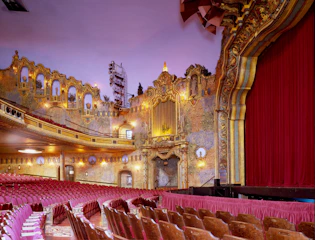 An ornate theater interior featuring elaborate architectural details and a large red curtain. The theater has rows of red upholstered seats, decorative arches, and intricate moldings. Several balconies with golden embellishments and full of light fixtures add to the grandeur.
