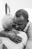 A heartfelt embrace between two people, one with short gray hair and the other with close-cropped hair. The setting appears intimate, suggesting comfort and warmth.