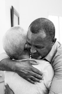 A heartfelt embrace between two people, one with short gray hair and the other with close-cropped hair. The setting appears intimate, suggesting comfort and warmth.