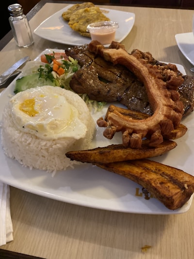 A plate of food includes a portion of white rice topped with a fried egg, alongside grilled meat and crispy fried pork skin. Accompaniments include fried plantains and a small side salad with cucumbers and carrots. In the background, there is a small cup of sauce and some fried plantains on another plate.