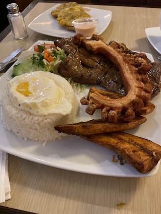 A plate of food includes a portion of white rice topped with a fried egg, alongside grilled meat and crispy fried pork skin. Accompaniments include fried plantains and a small side salad with cucumbers and carrots. In the background, there is a small cup of sauce and some fried plantains on another plate.