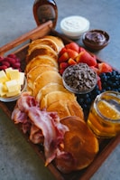 A wooden tray filled with a variety of breakfast items including pancakes, crispy bacon strips, a bowl of raspberries, a plate of butter cubes, a bowl of chocolate chips, fresh strawberries, blueberries, honey in a jar, and bowls of cream and chocolate spread. A bottle of maple syrup is placed in the background.