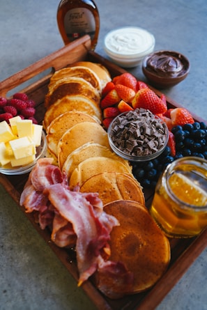 A wooden tray filled with a variety of breakfast items including pancakes, crispy bacon strips, a bowl of raspberries, a plate of butter cubes, a bowl of chocolate chips, fresh strawberries, blueberries, honey in a jar, and bowls of cream and chocolate spread. A bottle of maple syrup is placed in the background.