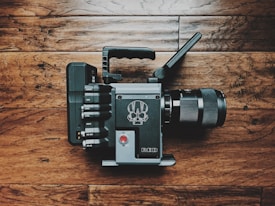 A professional video camera placed on a wooden floor, featuring a handle on top and several connection ports on the side. The camera body is sleek and modern, with a prominent red button and a distinctive logo design. The lens is large and prominent, indicating advanced functionality for filmmaking.