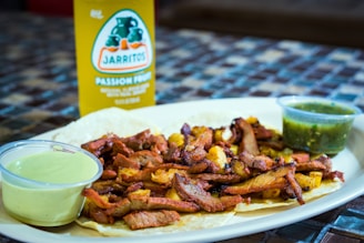 A plate of tacos filled with grilled meat and pineapple is accompanied by two small containers of sauce. Behind the plate, a bottle of Jarritos passion fruit soda is visible.