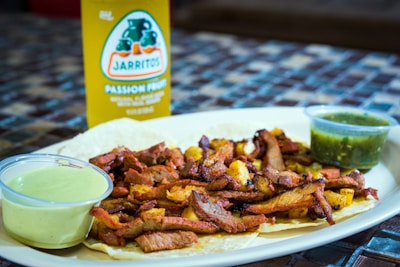 A plate of tacos filled with grilled meat and pineapple is accompanied by two small containers of sauce. Behind the plate, a bottle of Jarritos passion fruit soda is visible.