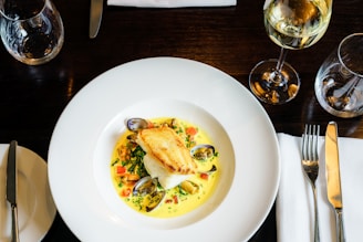 A dish featuring a piece of seared white fish placed on top of a bed of vegetables and shellfish in a creamy sauce. The meal is elegantly presented in a large white bowl with a neatly arranged dining setting including a knife, fork, and a glass of white wine.