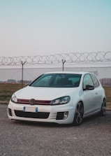 A white compact car is parked on a gravel surface in front of a chain-link fence topped with barbed wire. The sky above is clear, with a subtle gradient from light blue to pink. The car's design features sporty elements, including a sleek aerodynamic build and distinctive red accents on the grille.