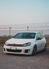 A white compact car is parked on a gravel surface in front of a chain-link fence topped with barbed wire. The sky above is clear, with a subtle gradient from light blue to pink. The car's design features sporty elements, including a sleek aerodynamic build and distinctive red accents on the grille.